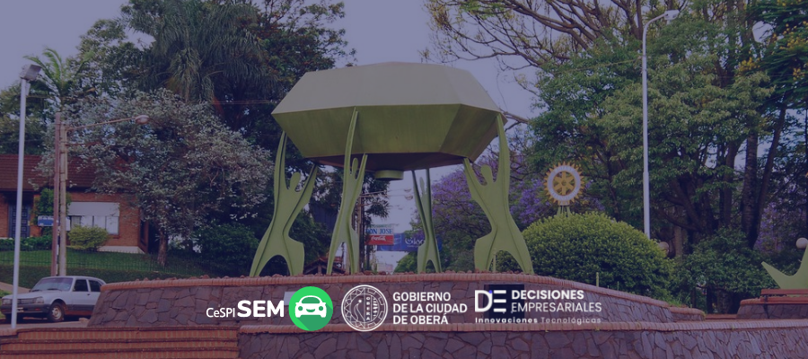 Monumento emblemático de Oberá con árboles y casas al fondo, junto a logos del sistema SEM, Gobierno de la Ciudad de Oberá y Decisiones Empresariales, representando la implementación del sistema de estacionamiento medido en la ciudad.
